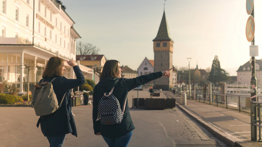 Zwei Frauen auf einem Tagesausflug nach Lindau, die eine Frau zeigt im Hafen auf etwas in der Ferne.