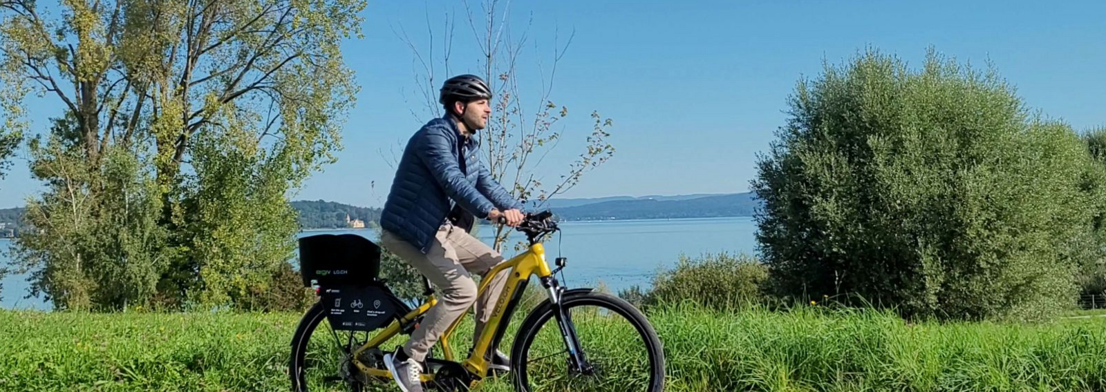 Mann fährt auf einem gelben Mietvelo von RegiVelo über einen Fahrradweg, im Hintergrund der Bodensee.