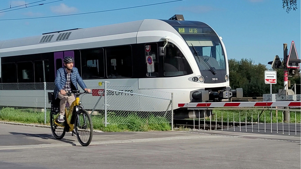Mann fährt entlang der Bahnlinie mit einem Mietvelo, im Hintergrund fährt ein Thurbo Zug bei geschlossener Barriere durch.